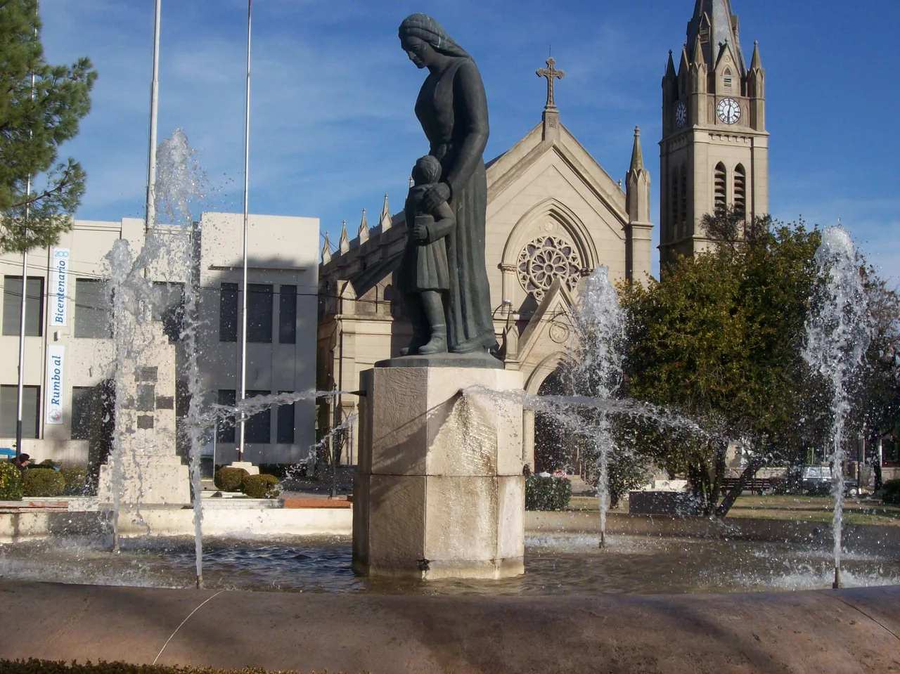 Plaza 1810 en la ciudad de Lobos, Provincia de Buenos Aires, Argentina. En primer plano se encuentra el Monumento a la Madre, al fondo, el Palacio Municipal de Lobos y la Parroquia Nuestra Señora del Carmen.