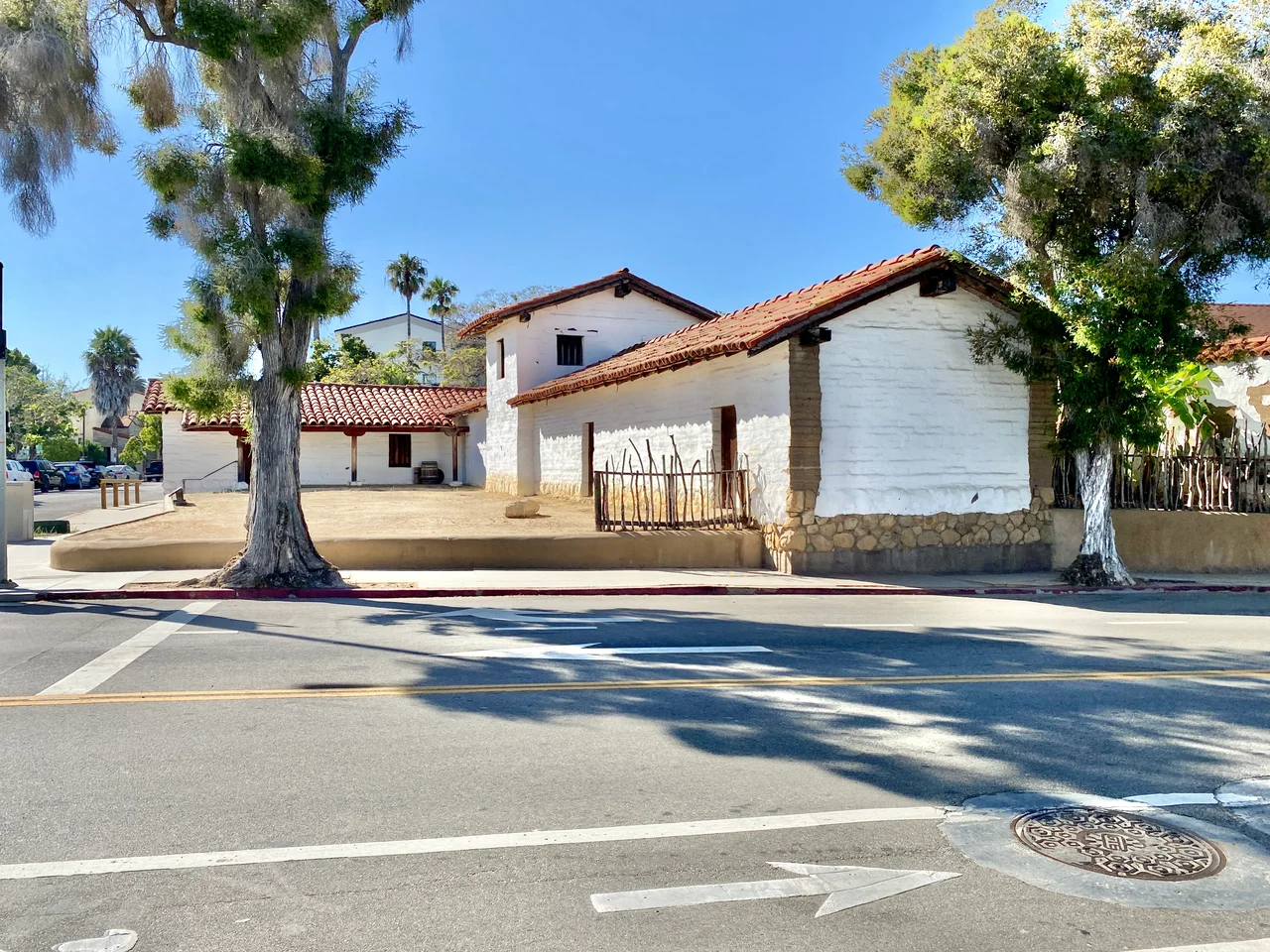 Originally built in 1782, this complex of buildings were constructed by the Spanish Colonial Authorities of California to serve as a presidio to protect the nearby Mission Santa Barbara and serve as a center of political influence in the surrounding region. The adobe buildings stood into the mid-19th Century, when they were slowly demolished and replaced by a neighborhood with roads platted through the middle of the presidio in the 1850s. By the mid-20th Century, only a single structure, the El Cuartel, which is the second-oldest existing building in California, remained standing, having been transformed into a private residence. The buildings were largely reconstructed after an archaeological investigation, with the first part of the complex being rebuilt between 1983 and 1985. The buildings consist of one-story adobe structures with red terra cotta tile roofs, exposed rafter tails, porches with wooden columns, and small window openings surrounding a central dirt courtyard, which has been partially restored, and a larger and taller chapel at the center of the western end of the courtyard, with a gabled roof, an arched entrance bay, and small tower with two bells. The buildings are divided by modern streets laid out after the Presidio had been largely demolished, with the ends of the reconstructed buildings revealing the interior structure of the presidio buildings where they are cut away along the streets. The original buildings in the complex were listed on the National Register of Historic Places in 1973. Today, the reconstructed Presidio serves as a historical museum operated by the Santa Barbara Trust for Historic Preservation.