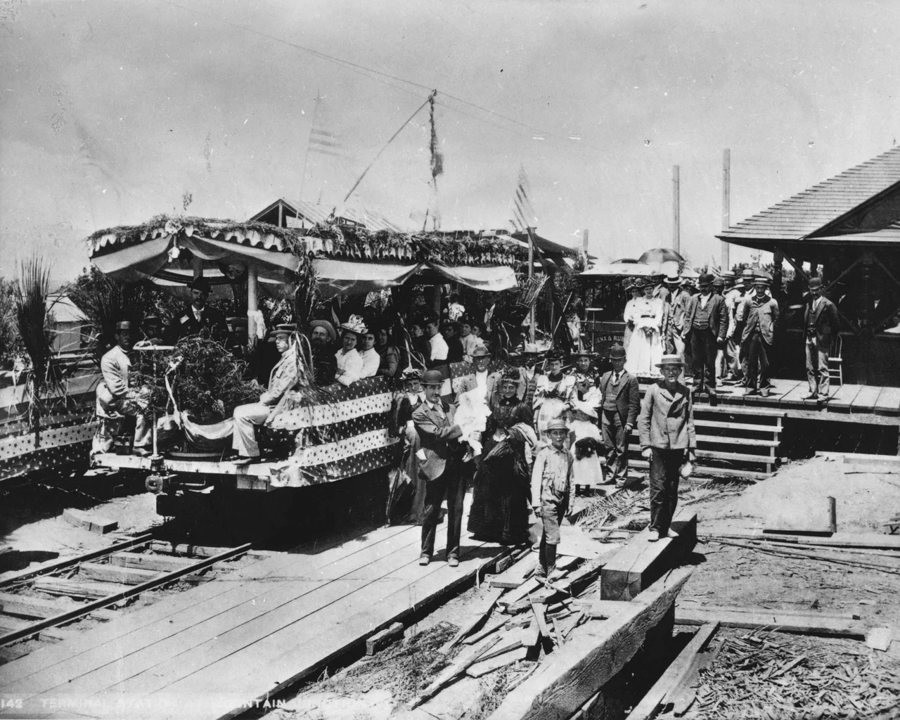 Photograph of the first Mount Lowe Railway car to leave Altadena for Rubio Canyon, July 4, 1893. A large group of finely-dressed people gather on the railroad platform looking towards the foreground. Many stand on the raised, wooden platform at right while others sit on a decorated railroad car at center. Two small boys stand on broken shards of wood in the foreground while flags wave in the background.