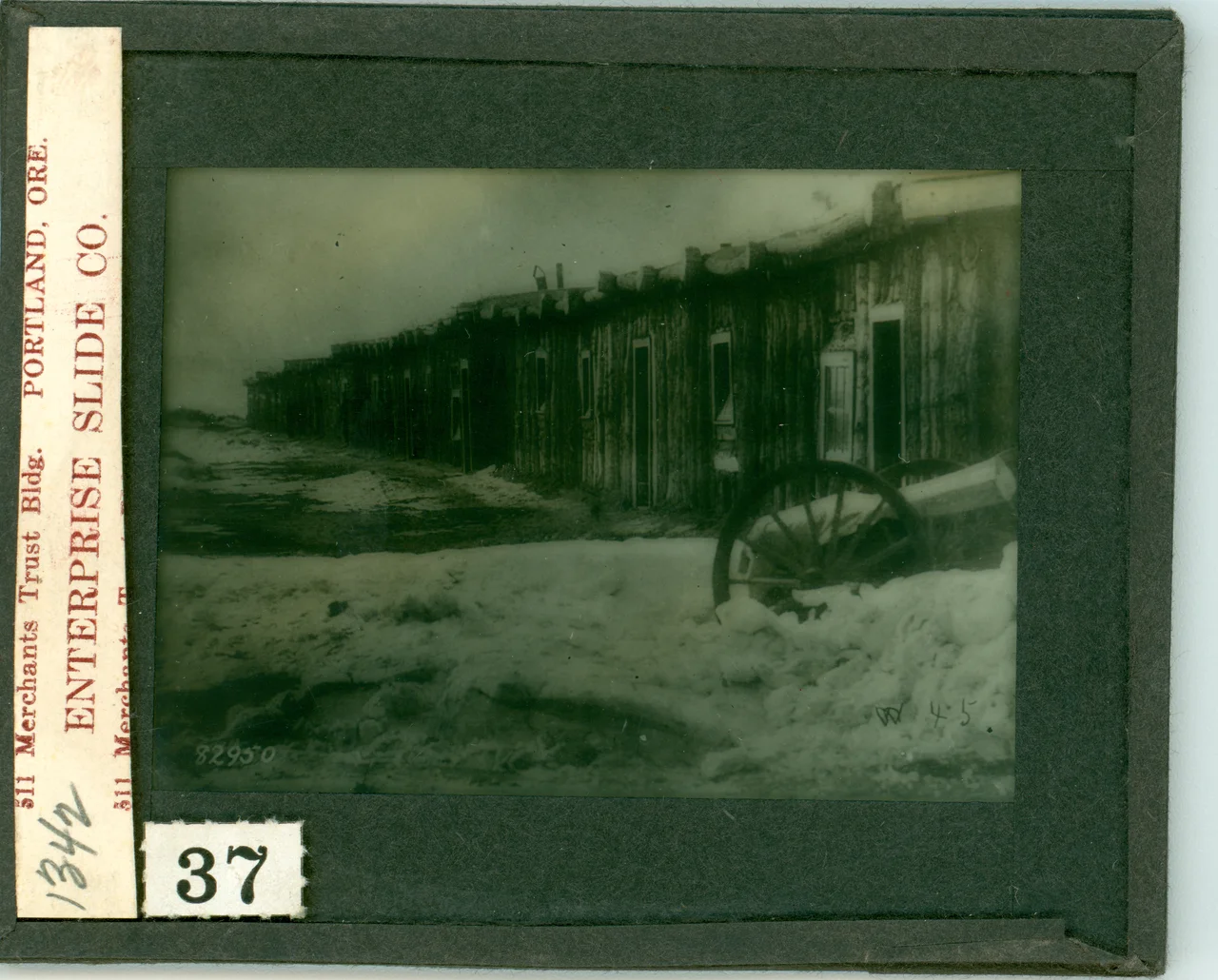LIBI_00030_01342; A lantern-slide (glass transparency), "A part of the cantonment at the Tongue River". A view of a row barrack buildings that later became part of Ft. Keogh in the snow. It was produced by an unknown photographer  in 1877. The number “82950” is printed in the left corner. A printed label on the left reads: “511 Merchants Trust Bldg. Portland, OR Enterprise Slide Co.” [Bowen No. 37; picture file &amp; negative]; transparency, latern-slide [photographic material];  Courtesy of the National Park Service, Little Bighorn Battlefield National Monument, LIBI_00030_01342, Unknown Photographer, "Row of Wooden Barracks That Would Become Part of Fort Keogh in the Snow," circa 1877
Keywords: little bighorn battlefield national monument