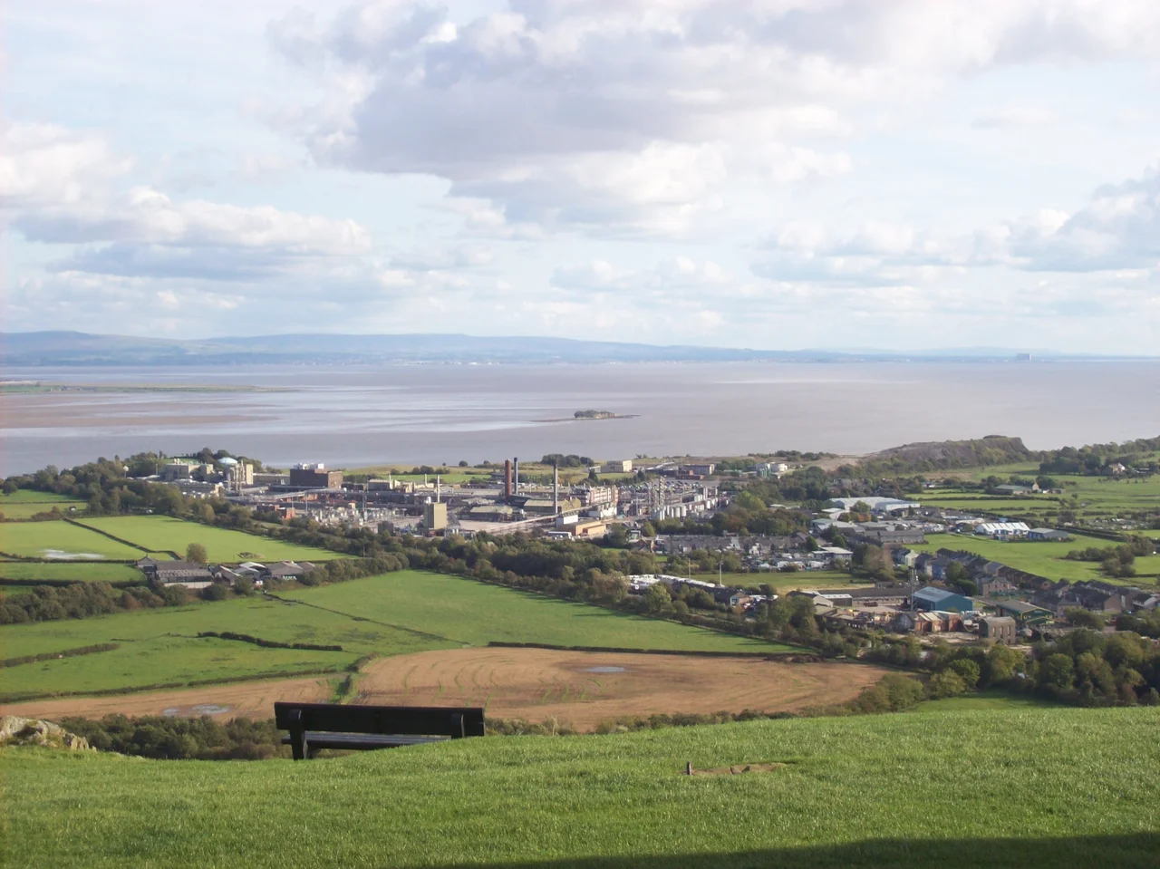 Morecambe Bay from Ulverston. Vantage point at the top of Hoad Hill.
In the immediate foreground is some agricultural land. On the other side of Ulverston Canal is the industrial area, which is a GlaxoWellcome factory. Along the canal's banks are some abandoned buildings, some houses, and some caravans. The River Leven reaches the ocean here, thus becoming the Leven estuary. The small island is Chapel Island. At the centre-far-right is a small rectangle, we call that Morecambe.