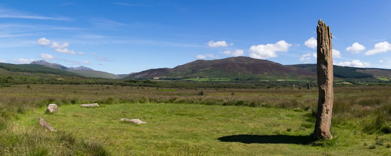 Circle 3 at Machrie Moor.
One remaining stone standing 4.3m tall. The stumps of three others remain. In the middle distance to the right are the three large stones of circle 2. In the far distance to the left are the mountains of the north of Arran, consisting of Beinn Tarsuinn and Goat Fell.