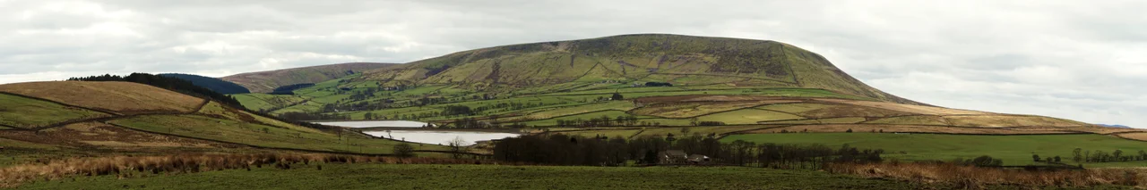 A panoramic image of Pendle Hill, Lancashire, showing the northeast slopes, known as the Big End, photographed from Wheathead Lane on the edge of Barley-with-Wheatley Booth. To the left are Aitken Wood and Black Moss Reservoirs. The footpath from Barley (not pictured) to the summit can be seen ascending from left to right. To the far right of the image, visible in the distance 8.25 miles away, is the radio mast on Waddington Fell.