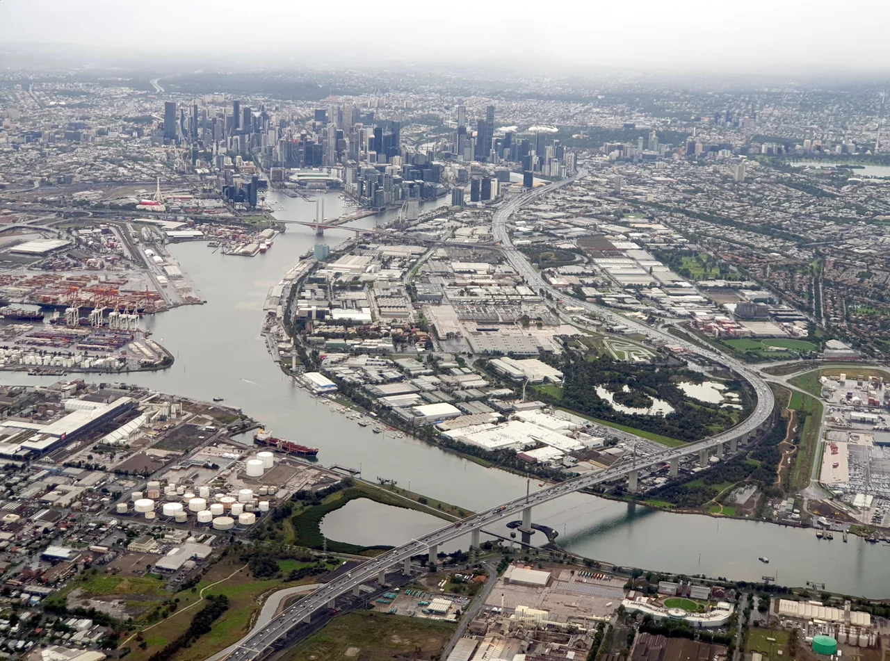 An aerial photograph looking east from near the Yarraville Oil Terminal (foreground left) along the Yarra River to Melbourne CBD, with the West Gate Bridge and Fishermans Bend prominently visible as well as Coode Island (left) and the start of Port Melbourne and Albert Park (right).