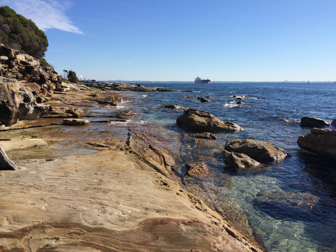 In this image, taken from Inscription Point on the South Head region of Kamay Botany Bay National Park, we look across Botany Bay, looking northwest in the direction of an outbound cargo ship, presumably originating from Port Botany. In the foreground, weathered and eroded rocks sit idle in the water and are constantly splashed by the occasional wave.