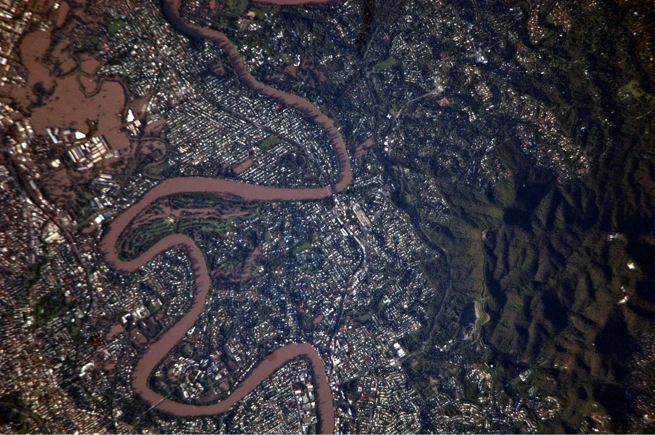 This detailed satellite photograph illustrates flooding in metropolitan suburbs of the Brisbane, Queensland, Australia. Please note that the top of the photo is oriented approximately to the South-West and not to the North. The image, taken by astronauts on board the International Space Station, highlights several suburbs along the Brisbane River in the southern part of the Brisbane metropolitan area. The light-coloured rooftops of residences and other structures contrast sharply with green vegetation and brown, sediment laden flood-waters. Most visible low-lying areas are inundated by flood-water, perhaps the most striking being Rocklea at image upper left. The suburb of Yeronga (image lower left) also has evident regions of flooding, as does a park and golf course located along a bend in the Brisbane River to the south of St. Lucia (image center). Flooding becomes less apparent near the higher elevations of Mt. Coot-Tha at image right.