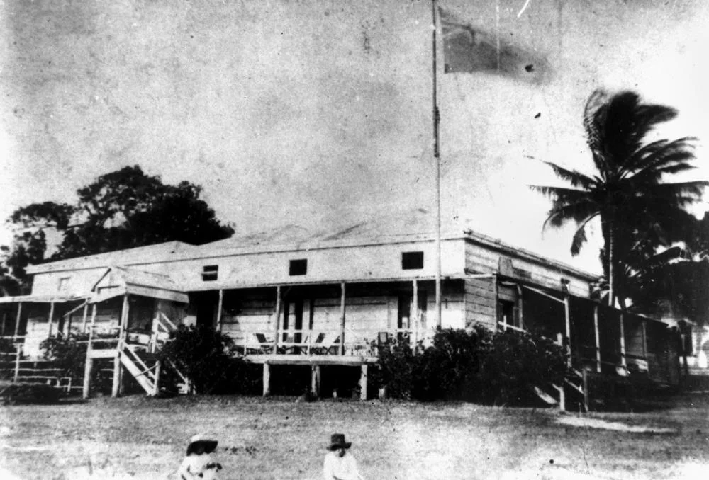 Government House, the home of Frank Jardine in Somerset, Cape York, Queensland. The home is a timber structure with external weatherboards and a plantation style iron roof. Some deck chairs are on the verandah and a flag is flying from the flag pole in front of the house. Two small children in the foreground may be Frank Jardine's.