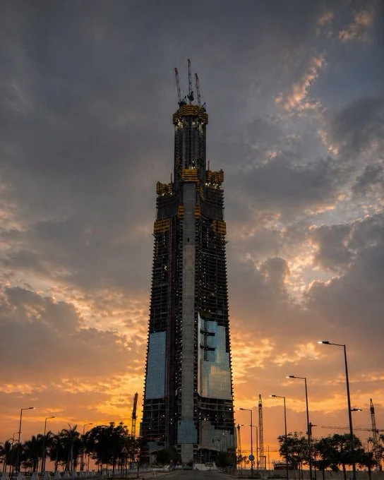 A 2026 photo of Jeddah Tower in Jeddah under construction after reaching the 100th floor, with cranes visible at the top and sunset light reflecting on the glass façade against a cloudy evening sky.