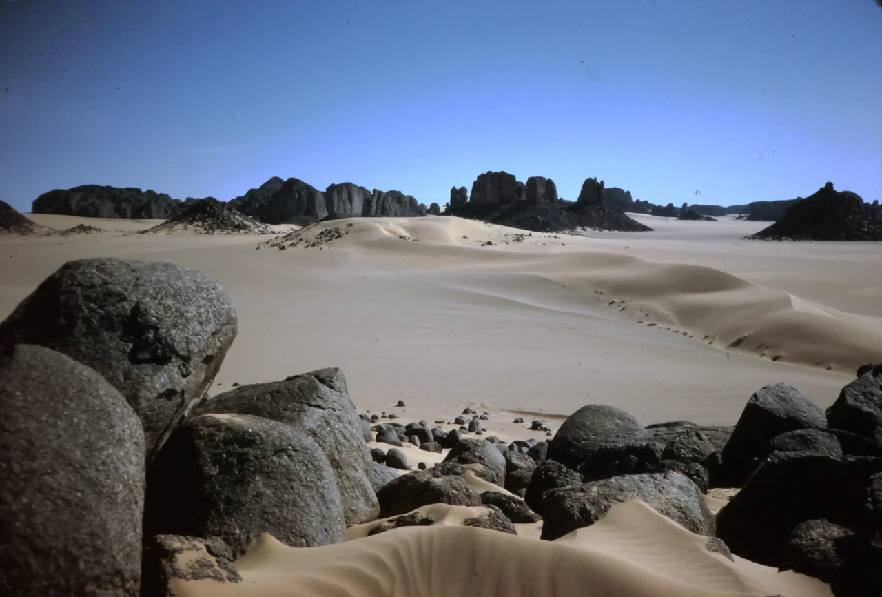 Location: On the southeastern edge of Tassili n'Ajjer, northwest of Djanet
The prevailing wind in the Sahara is the northeast trade wind (Harmattan). During the early Quaternary period, there were times when it blew much stronger than it does today. The sand in this picture was probably blown at that time from the Erg Ubari and Erg Murzouk areas in southwestern Libya across the gently ascending plateau of the Tassili n'Ajjer and deposited here, in the lee behind the mountain ridge.

The rounded boulders in the foreground are the result of insolation weathering. The large temperature differences between day and night build up tension in the rock, especially on protruding parts. These splinter off, and as the boulder becomes smaller and smaller, its shape becomes more and more rounded.