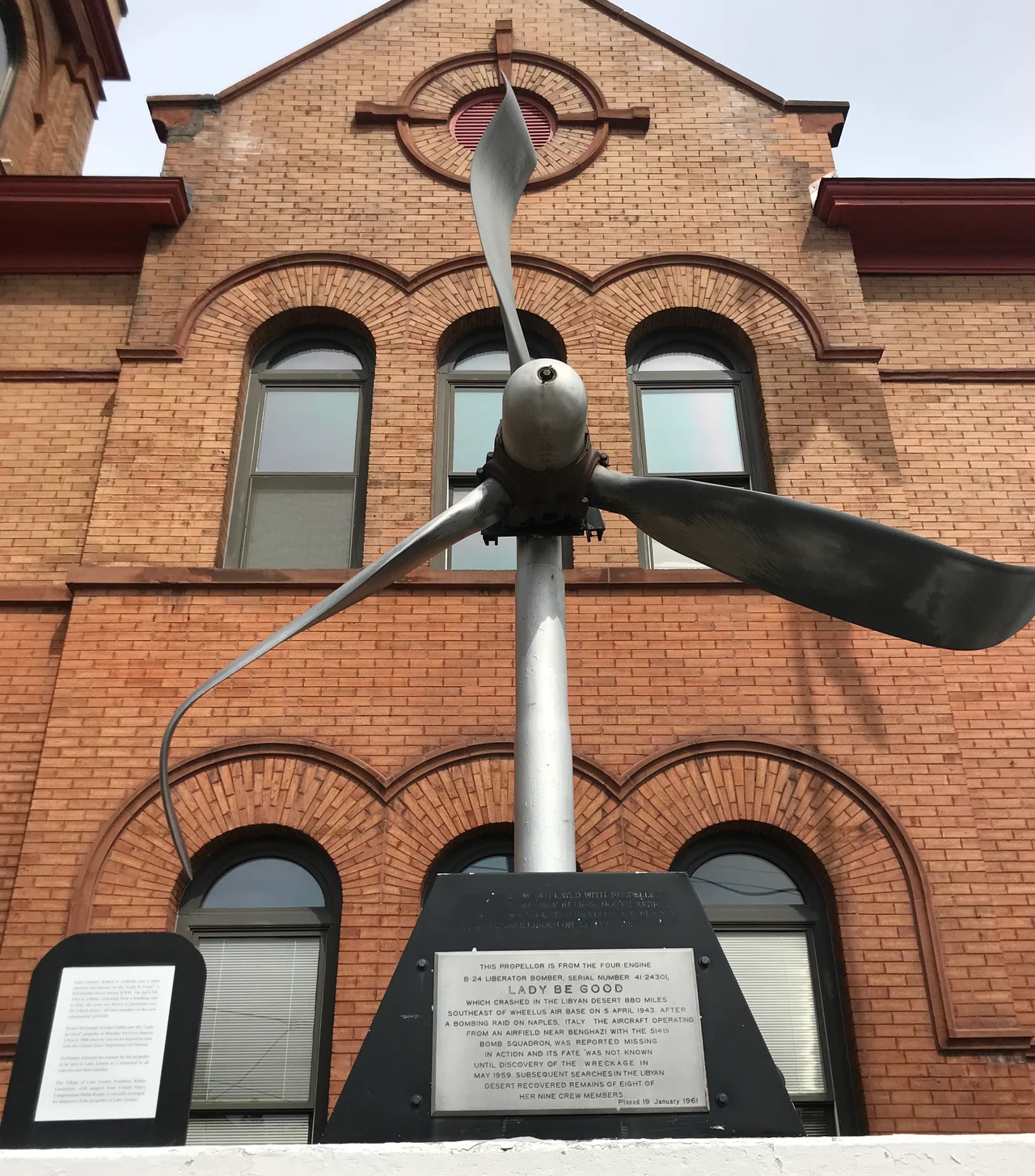 A damaged propeller from the Lady Be Good displayed in Lake Linden, Michigan. The text of the plaque reads “This propeller is from the four-engine B-24 Liberator bomber, serial number 41-24301, Lady Be Good which crashed in the Libyan desert 880 miles southeast of Wheelus Air Base on 5 April 1943, after a bombing raid on Naples, Italy. The aircraft operating from an airfield near Benghazi with the 514th Bomb Squadron, was reported missing in action and its fate was not known until discovery of the wreckage in May 1959. Subsequent searches in the Libyan desert recovered remains of eight of her nine crew members. Placed 19 January 1961.” A placard to the side reads “Lake Linden’s Robert E. LaMotte was a radio operator and gunner on the ‘Lady be Good’ a B25 bomber active during WWII. On April 5th, 1943 at 2:00 a.m., returning from a bombing raid to Italy, the crew was forced to parachute over the Libyan desert. All nine members of the crew subsequently perished. / Octave DuTemple of Lake Linden saw the ‘Lady Be Good’ propeller at Wheelus Air Force Base in Libya in 1968 when he was on an inspection tour with the United States Department of Defense. / DuTemple initiated the request for the propeller to be sent to Lake Linden as a memorial to all veterans and their families. / The Village of Lake Linden President Walter Laverdiere [1923–2014], with support from United States Congressman Philip Ruppe, eventually arranged for the shipment of the propeller to Lake Linden.”