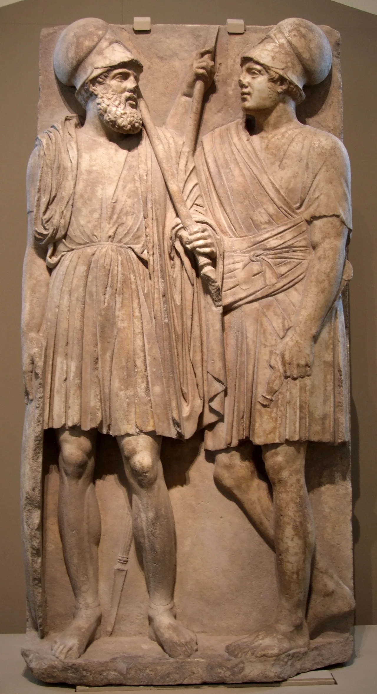 Stele with two warriors of the Bosporan Kingdom. From Taman peninsula (Yubileynoe), Southern Russia, 3rd quarter of the 4th century BC. Marble, Pushkin museum
The marble relief was found in 1985. It is 1.88 m high, above 0.85 m and below 0.94 m wide. The plate is 0.25 m thick, the relief protruding 0.12 m. Also the whole surface of the plate is worked – the shield of the older warrior is reaching to the very margin of the stone. Two soldiers are represented, gazing each other. The bearded soldier has all attributes of a hoplite; the younger is only holding a spear.